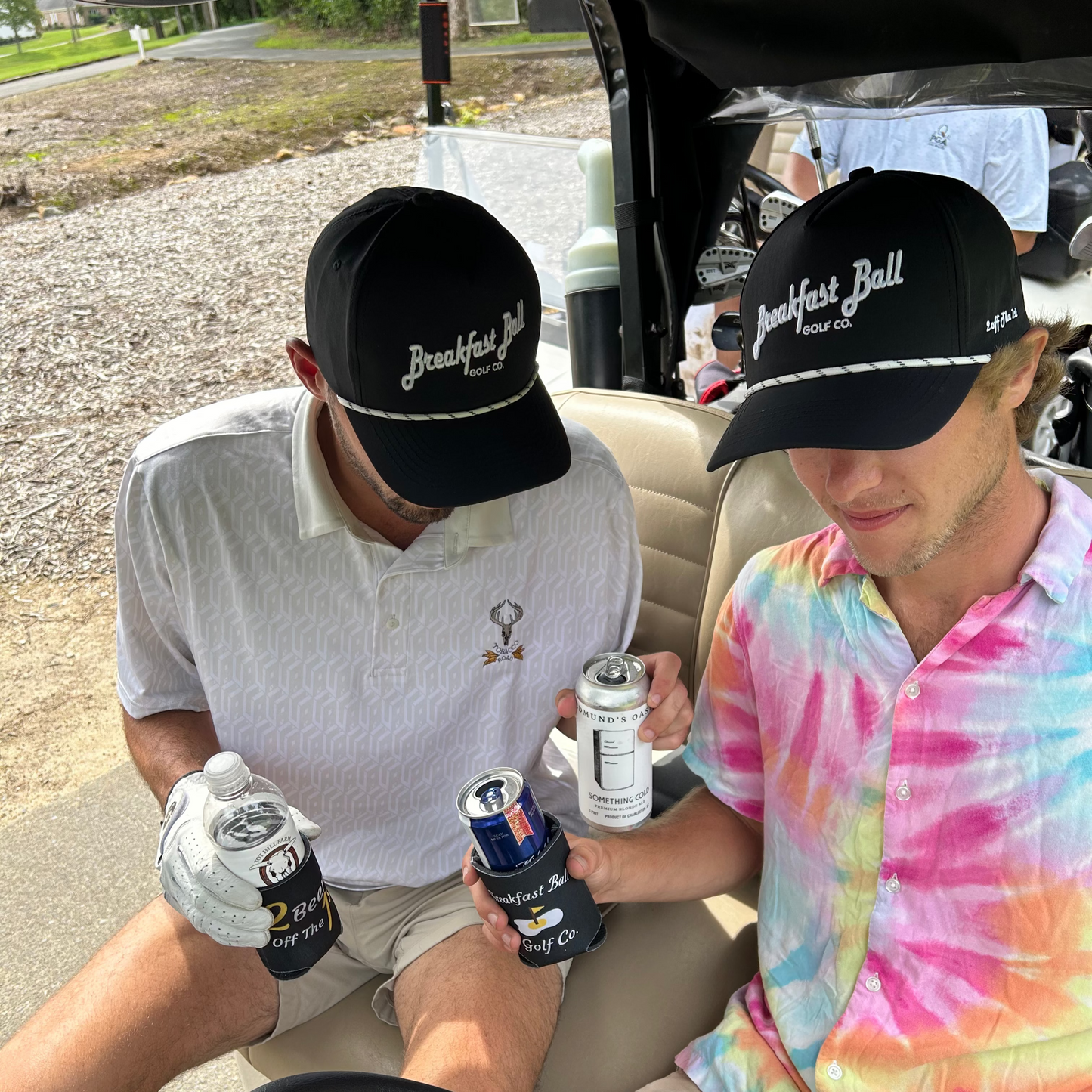Two people in a golf cart wearing 'Breakfast Ball' hats, holding drinks.